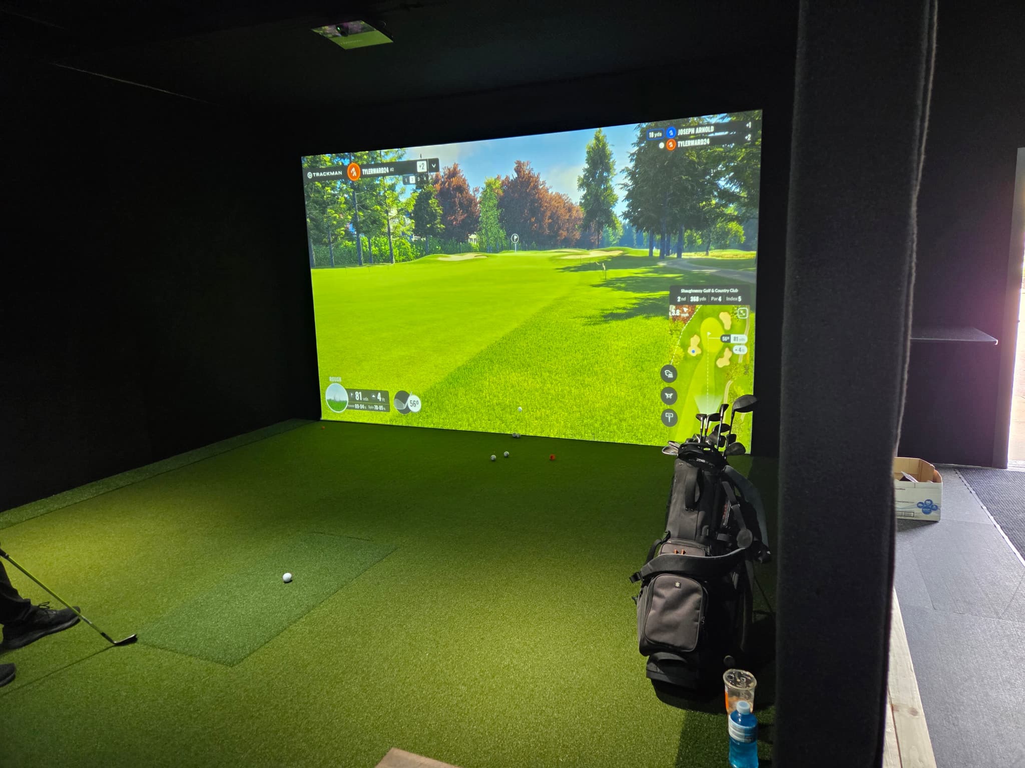 Simulator bay with a ceiling-mounted TrackMan iO, a projected golf course, a golfer’s feet and club at left, and a golf bag at right